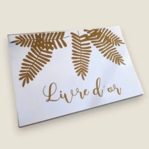Un livre d'or de mariage rectangulaire au format paysage avec une couverture blanche épurée, illustré de feuilles de fougère dorées suspendues sur la partie supérieure et de l'inscription calligraphiée dorée "Livre d'or".