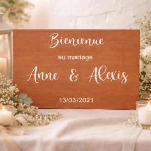 Un panneau rectangulaire en bois teinté marron chaud posé à l'horizontale sur une nappe blanche, au centre d'une décoration de mariage romantique. La pancarte affiche l'inscription en lettres blanches "Bienvenue" en calligraphie cursive, suivie de "au mariage" en lettres scriptes, des prénoms des mariés "Anne & Alexis" en grande écriture manuscrite, et de la date "13/03/2021" à la base. L'ensemble est entouré de bouquets de roses blanches, de gypsophile, d'une lanterne blanche et de bougies allumées dans des photophores.