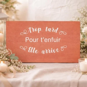 Un panneau rectangulaire en bois teinté d'une couleur chaleureuse terre cuite ou terracotta, posé sur une nappe blanche dans un décor romantique entouré de roses blanches, de gypsophile et de bougies allumées. La pancarte affiche l'inscription humoristique en blanc "Trop tard" en calligraphie cursive, suivi de "Pour t'enfuir" en lettres bâtons épurées, et "Elle arrive" en bas, le tout encadré par de délicates arabesques et volutes blanches.