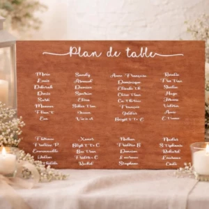 Un grand panneau rectangulaire en bois brut teinté marron chaud, posé à l'horizontale sur une nappe blanche dans une ambiance romantique. Le panneau affiche le titre "Plan de table" écrit en lettres cursives blanches élégantes avec de fines lignes de part et d'autre. En dessous, la liste des invités est organisée et écrite en blanc, répartie sur quatre colonnes distinctes. Le décor environnant comprend des roses blanches, des nuages de gypsophile, une lanterne blanche en bois et des bougies allumées dans de petits photophores en verre.