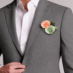 Boutonnière de mariage classique pour homme, composée d'une rose en papier couleur pêche, d'une rose vert sauge et de feuillages verts discrets, portée sur le revers d'un costume gris.