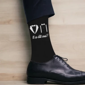 Plan rapproché sur une cheville d'homme portant une chaussette noire à motifs blancs représentant une veste de costume et des bretelles avec l'inscription cursive "Il a dit oui !", associée à une chaussure Richelieu noire.