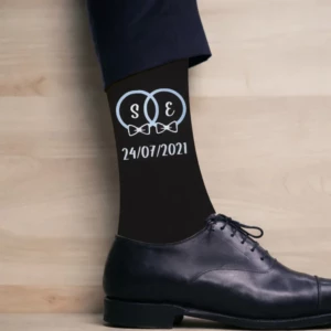Vue rapprochée d'une cheville d'homme avec une chaussette noire personnalisée affichant deux alliances entrelacées contenant les initiales S et E avec des nœuds papillons, surmontant la date blanche 24/07/2021, portée avec une chaussure Richelieu en cuir noir.