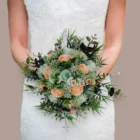 Bouquet de mariée évasé et champêtre composé de roses en papier couleur corail, de feuillage vert et de lettres en bois épelant le mot YES, tenu par une mariée en robe blanche.