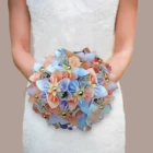 Vue centrée d'une mariée tenant un bouquet rond original en origami, composé de fleurs en papier aux tons bleu pastel, corail et pages de livre imprimées, avec des perles dorées au centre de chaque fleur.