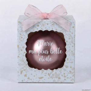Boîte cadeau blanche à motifs dorés et étoilés avec un nœud rose, contenant une boule de Noël rose nacrée avec l'inscription "Marie, ma plus belle étoile".
