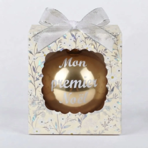 Boîte cadeau beige à motifs floraux et argentés avec un nœud argenté, contenant une boule de Noël dorée brillante avec l'inscription "Mon premier Noël".