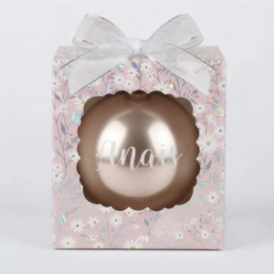 Boîte cadeau rose à motifs de marguerites blanches avec un nœud argenté, contenant une boule de Noël cuivrée ou rose dorée avec le prénom "Anais".