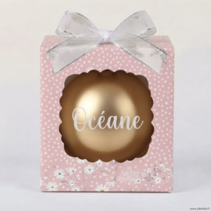Boîte cadeau rose à pois blancs et détails floraux avec un nœud argenté, contenant une boule de Noël dorée avec le prénom "Océane".