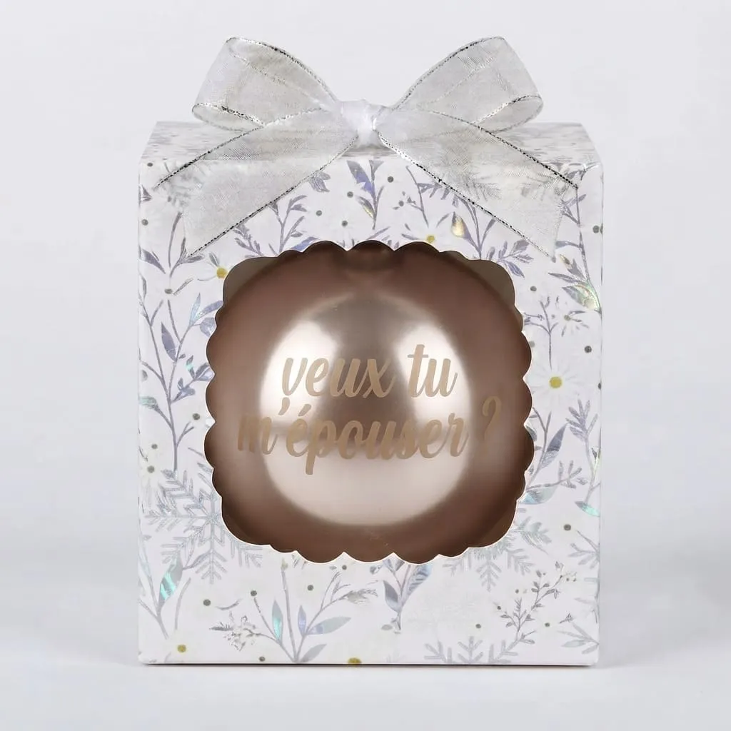 boule-de-noel-veux-tu-m-epouser Boîte cadeau blanche à motifs végétaux et flocons argentés avec un nœud argenté, contenant une boule de Noël beige nacrée avec le texte "veux tu m'épouser ?".