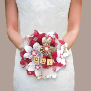 Bouquet de mariée rond fait main avec des fleurs en origami blanches, rouges et en papier livre, orné de plumes, de perles et de tuiles en bois formant le mot OUI.