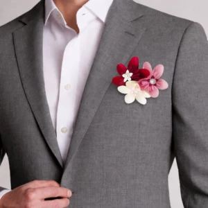 Boutonnière de mariage au design minimaliste composée de trois fleurs en papier lisse aux nuances rouge bordeaux, rose et blanc crème avec de petits cœurs perlés, fixée sur le revers d'une veste grise.