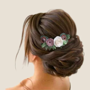 Un chignon de mariée tressé et élégant, de couleur marron, orné d'un peigne à cheveux floral composé de roses en tissu vieux rose, bordeaux et blanc, avec des feuilles d'eucalyptus artificielles.