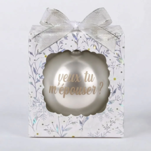 Boîte cadeau fleurie et enneigée avec une boule de Noël blanche nacrée portant l'inscription Veux-tu m'épouser ?