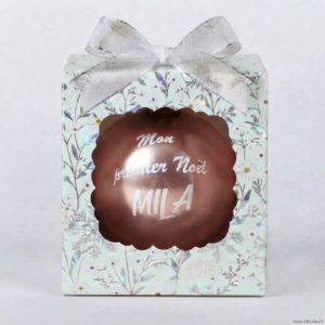 Boîte cadeau verte à motifs floraux avec un nœud argenté, contenant une boule de Noël marron personnalisée avec le texte "Mon premier Noël MILA".