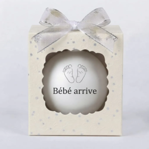 Boîte cadeau beige avec un nœud argenté et une boule de Noël blanche affichant des empreintes de pieds de bébé et le texte Bébé arrive