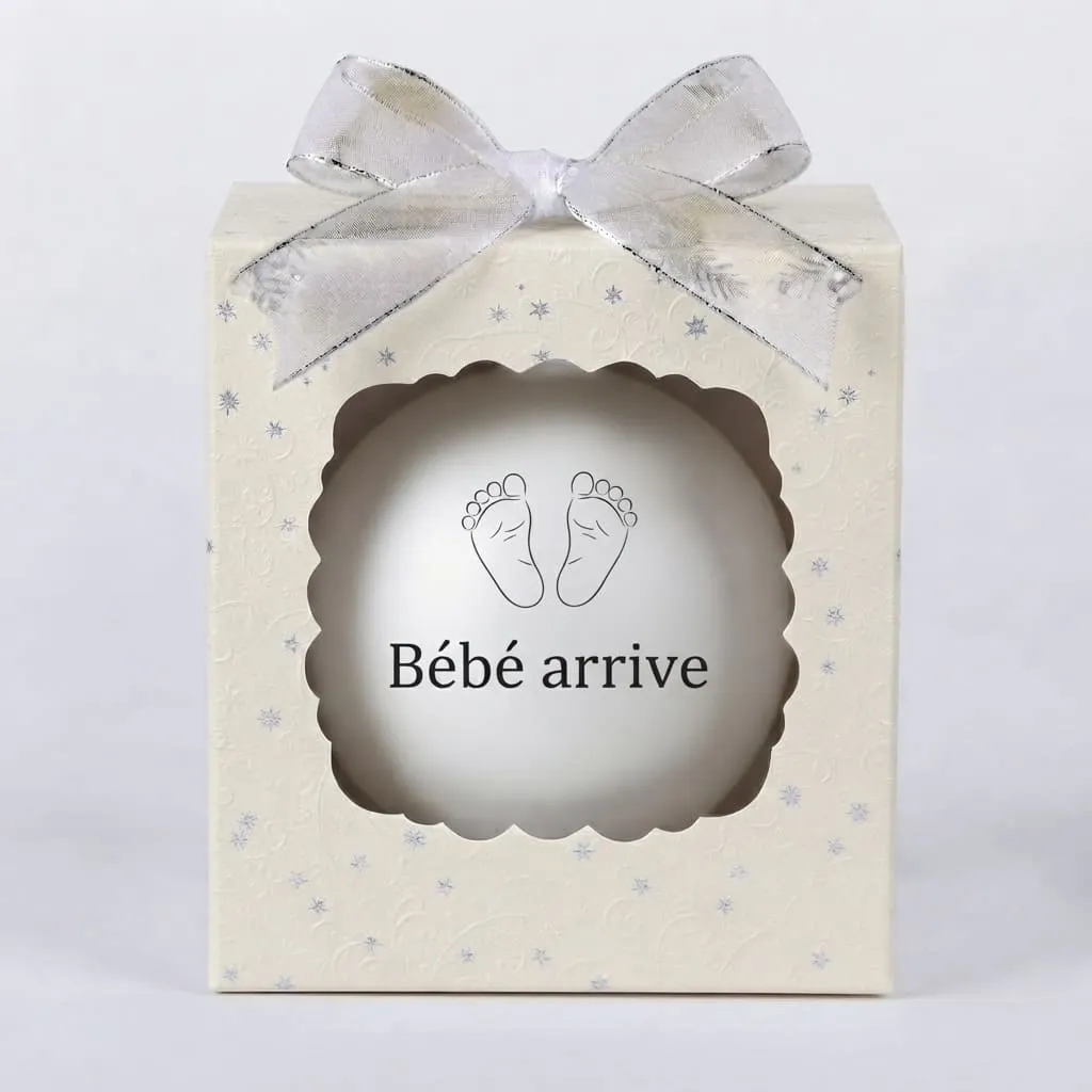 boule de noel personnalisee beb arrive Boîte cadeau beige avec un nœud argenté et une boule de Noël blanche affichant des empreintes de pieds de bébé et le texte Bébé arrive