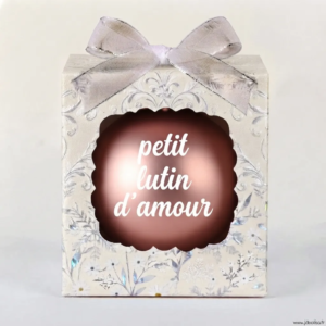 Boîte cadeau beige à motifs floraux et argentés avec un nœud transparent, contenant une boule de Noël marron/cuivrée avec l'inscription "petit lutin d'amour".