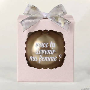 Boîte cadeau rose à motifs discrets avec un nœud argenté, contenant une boule de Noël dorée avec l'inscription "veux tu devenir ma femme ?".