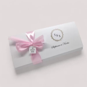 Un faire-part de mariage rectangulaire au format pochette allongée blanche, orné à droite du monogramme S & N dans une couronne de feuillage dorée et des prénoms Stéphanie et Nicolas écrits en lettres cursives, fermé sur la gauche par un ruban en satin rose noué avec une petite étiquette carrée indiquant la date 18 Juillet 2020.