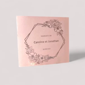 Un faire-part de mariage carré en papier rose poudré délicatement texturé, orné d'un cadre géométrique en forme d'hexagone asymétrique rehaussé de compositions florales de roses en dorure à chaud brillante, entourant l'inscription "Mariage de Caroline et Jonathan" et la date "30/08/2024".