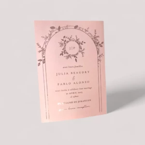 Un faire-part de mariage rectangulaire vertical en papier rose poudré texturé, illustré d'une arche double surmontée de feuillages et fleurs en dorure à chaud brillante, encadrant le monogramme J P, les prénoms Julia Beaudry & Pablo Alonso, et l'invitation à l'église de Jurançon.