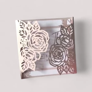 Un faire-part de mariage carré avec une pochette rose gold métallique ouverte au centre, dotée d'une découpe laser fine en forme de roses et de feuilles style dentelle, laissant entrevoir le carton d'invitation blanc intérieur avec des détails imprimés.