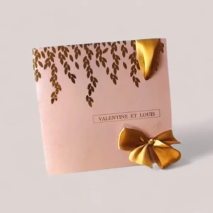 n faire-part de mariage carré en papier rose poudré haut de gamme, orné sur la partie supérieure d'une cascade poétique de petites feuilles en dorure à chaud dorée, affichant un encadré central avec les prénoms "VALENTINE ET LOUIS", et agrémenté d'un ruban de satin doré brillant noué en bas à droite.