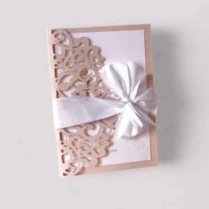Un faire-part de mariage rectangulaire vertical avec une pochette rose poudré découpée au laser style dentelle, laissant apparaître une carte d'invitation blanche à l'intérieur, le tout enveloppé d'un large ruban blanc satiné noué en un grand nœud papillon.