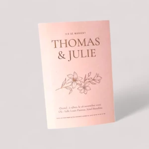 Un faire-part de mariage rectangulaire vertical en papier rose poudré de style minimaliste, affichant en haut la mention "Ils se marient", suivie des prénoms "Thomas & Julie" et d'une illustration de fleurs de lys délicates imprimées en dorure à chaud or rose brillante, avec les détails pratiques de la cérémonie inscrits en bas.