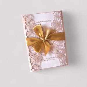Un faire-part de mariage rectangulaire vertical composé d'une pochette rose poudré découpée au laser avec des motifs d'arabesques style dentelle, s'ouvrant au centre pour laisser apparaître un carton d'invitation blanc, le tout encerclé par un large ruban de satin doré noué en un magnifique nœud papillon.