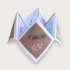 Un faire-part de mariage ludique plié en forme de cocotte en papier (origami), affichant sur un volet de couverture un fond kraft brun avec la date "07 juillet 2024" et une délicate illustration blanche de fleurs, entouré de volets intérieurs aux teintes pastel mauve et bleu ciel avec les inscriptions "FETE", "la vie" et "Le vin".