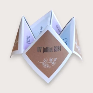 Un faire-part de mariage ludique plié en forme de cocotte en papier (origami), affichant sur un volet de couverture un fond kraft brun avec la date "07 juillet 2024" et une délicate illustration blanche de fleurs, entouré de volets intérieurs aux teintes pastel mauve et bleu ciel avec les inscriptions "FETE", "la vie" et "Le vin".