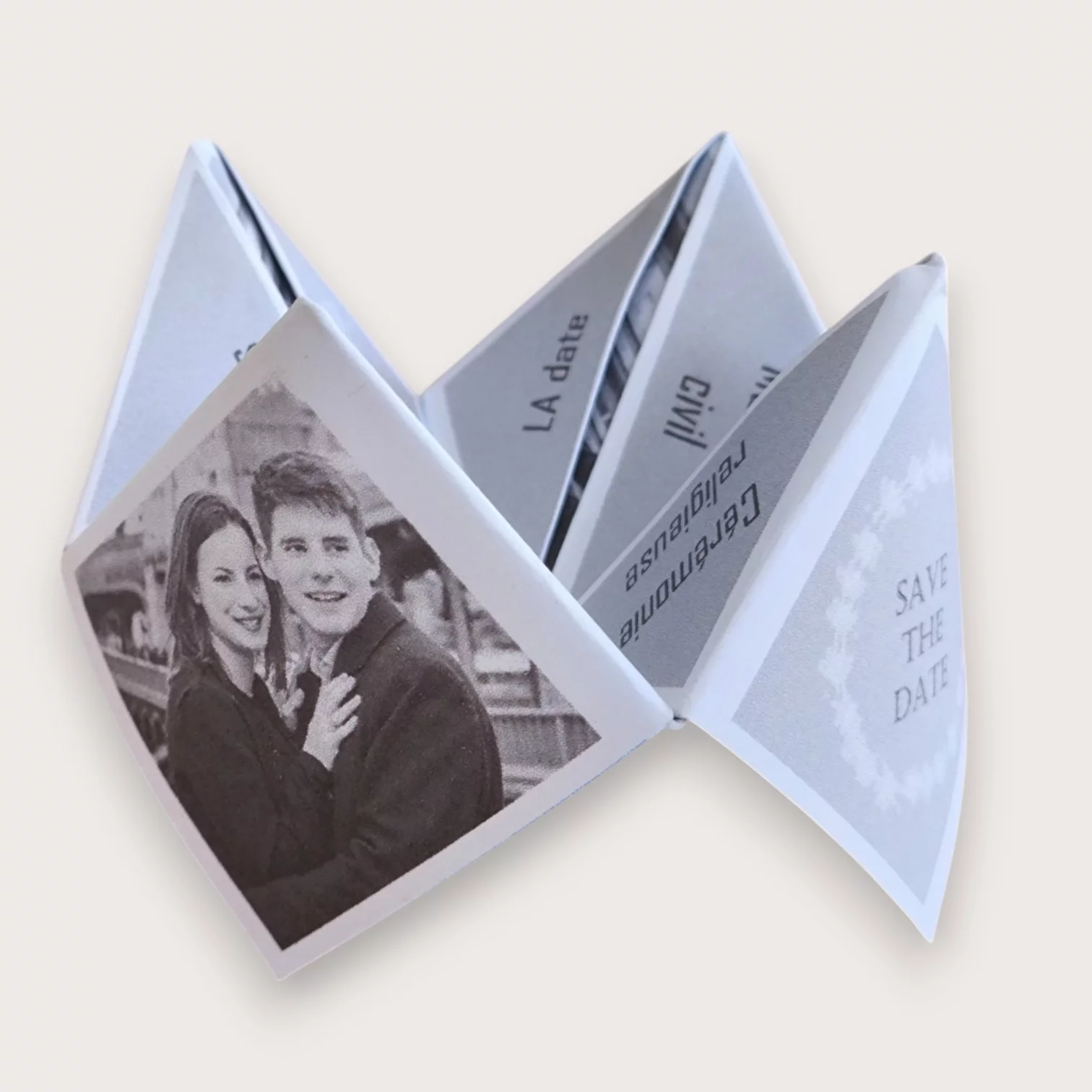 Un faire-part de mariage original plié sous forme de cocotte en papier blanc (origami), affichant sur un volet extérieur une photo en noir et blanc d'un jeune couple souriant, et sur le volet adjacent l'inscription "SAVE THE DATE" entourée d'une couronne végétale blanche sur fond gris, laissant entrevoir à l'intérieur les mentions "LA date", "civil" et "Cérémonie religieuse".