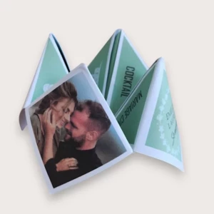 Un faire-part de mariage original plié en forme de cocotte en papier (origami) couleur vert sauge et blanc, affichant sur un volet extérieur une photo couleur romantique d'un couple complice, et sur le volet droit une couronne végétale entourant les prénoms des mariés, laissant entrevoir à l'intérieur les inscriptions "COCKTAIL" et "MARIAGE CIVIL".