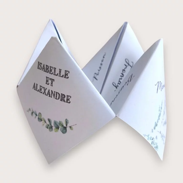 Menu de mariage original en cocotte en papier – Style Champêtre et Eucalyptus Un menu de mariage original plié en forme de cocotte en papier blanc (origami), affichant sur un volet extérieur les prénoms "ISABELLE ET ALEXANDRE" en lettres capitales grises au-dessus d'une délicate guirlande de feuilles d'eucalyptus, et laissant entrevoir à l'intérieur les catégories écrites à la main comme "Poisson" et "Fromage".