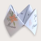 Un menu de mariage ludique plié sous forme de cocotte en papier blanc (origami), présentant sur un rabat extérieur le mot "Menu" écrit en calligraphie noire surmonté d'une fleur couleur pêche et de feuilles vertes, et sur le rabat opposé les prénoms "Julie et Christophe", révélant à l'intérieur les sections "Plat", "Entrée" et "Dessert".