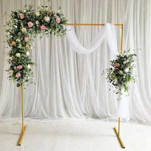 Une arche de mariage rectangulaire en métal doré, décorée d'une grande composition florale en haut à gauche et d'une plus petite à droite, avec des roses roses et blanches et du feuillage d'eucalyptus. Un voilage en mousseline blanche drape la barre transversale, devant un rideau de fond blanc plissé. L'arche est posée sur un sol blanc uni.
