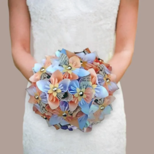 Vue centrée d'une mariée tenant un bouquet rond original en origami, composé de fleurs en papier aux tons bleu pastel, corail et pages de livre imprimées, avec des perles dorées au centre de chaque fleur.
