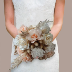 Bouquet de mariée composé de roses éternelles en satin blanc, de fleurs en papier crépon corail et de touches de fleurs séchées rose gold, tenu par une mariée en robe blanche.