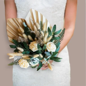 Bouquet de mariée moderne et éternel composé de roses en satin ivoire, de fleurs en papier vert sauge, de plumeaux de lagurus séchés, de feuillage de fougère et de feuilles de palmier séchées peintes en doré, tenu à deux mains par une mariée.