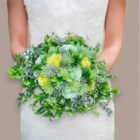 Mariée tenant un bouquet de fleurs évasé et champêtre, composé de roses éternelles en papier jaune vif et vert menthe, mélangées à d'abondants feuillages verts et plantes succulentes artificielles.