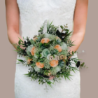 Bouquet de mariée évasé et champêtre composé de roses en papier couleur corail, de feuillage vert et de lettres en bois épelant le mot YES, tenu par une mariée en robe blanche.
