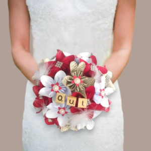 Bouquet de mariée rond fait main avec des fleurs en origami blanches, rouges et en papier livre, orné de plumes, de perles et de tuiles en bois formant le mot OUI.