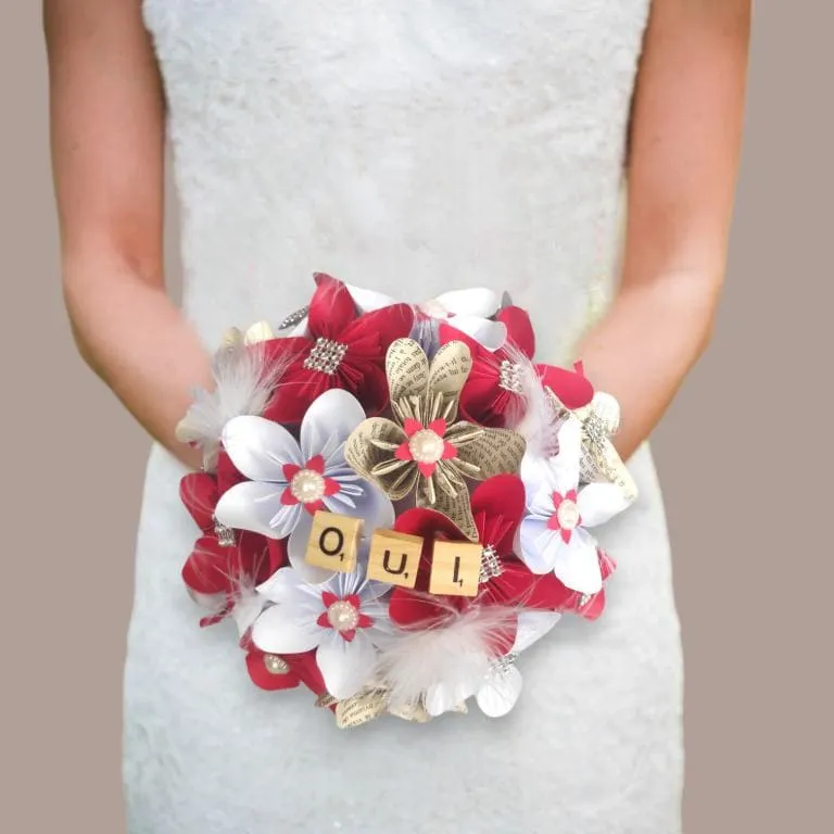 bouquet-mariee-rouge-blanc-fleurs-papier-idealisa Bouquet de mariée rond fait main avec des fleurs en origami blanches, rouges et en papier livre, orné de plumes, de perles et de tuiles en bois formant le mot OUI.