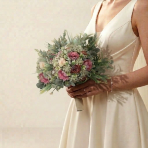 Bouquet de mariée bohémien rond composé de roses mauves, fleurs en papier et feuillage d'eucalyptus tenu par une mariée en robe ivoire.